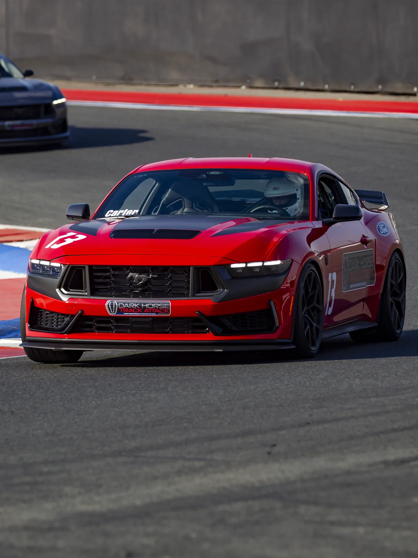 
A vibrant red Ford Mustang Dark Horse race car, with a number 13 on its side, is shown driving on a racetrack. 