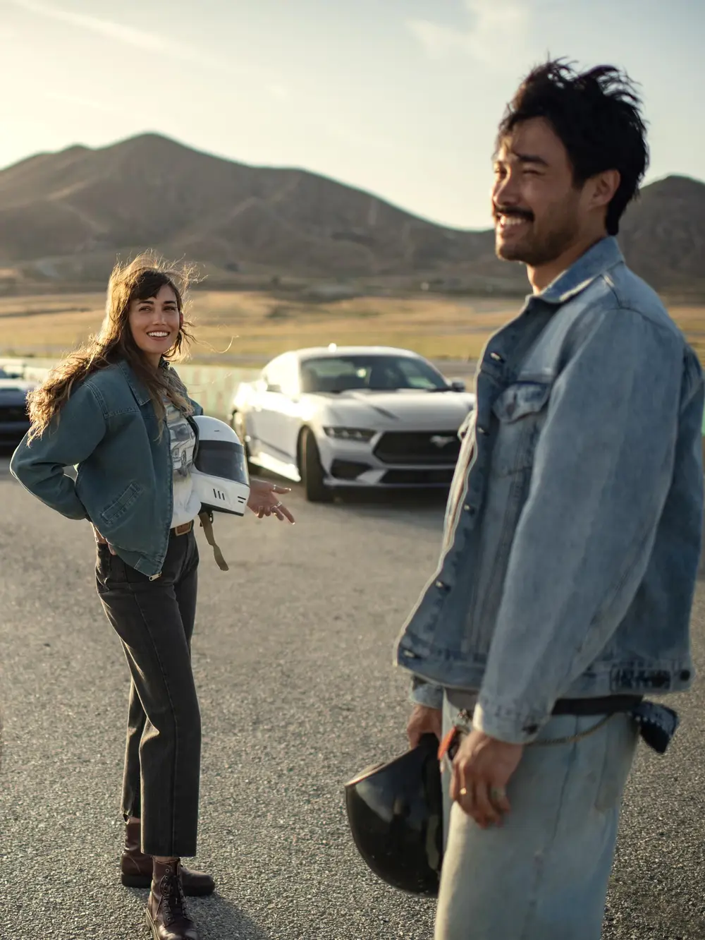  smiling man and woman stand on a racetrack with a white Ford Mustang in the background. 