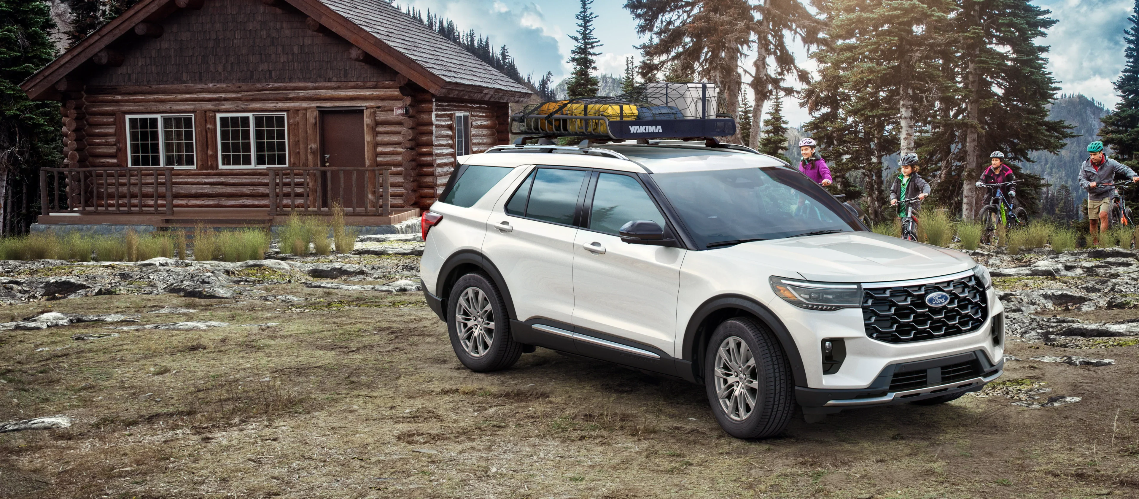 A white Ford Explorer SUV with a roof rack is parked in front of a log cabin, with a family on mountain bikes in the background.