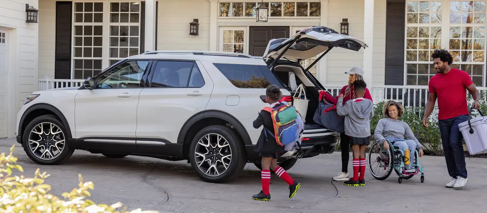 
A family is loading luggage into the back of a white Ford Explorer SUV parked in a driveway. A child in a wheelchair is smiling beside the open trunk.