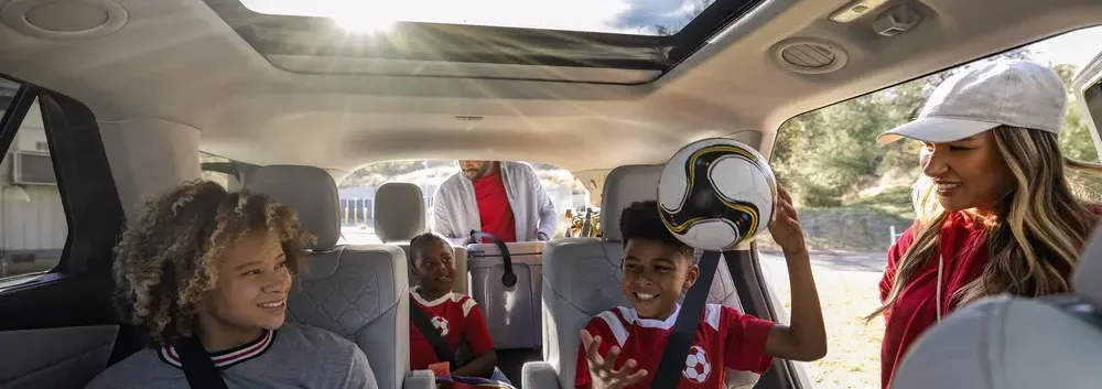 
A family is loading a Ford Explorer SUV, with two kids in the back seats and a man and woman standing near the open trunk. One of the kids is holding a soccer ball.