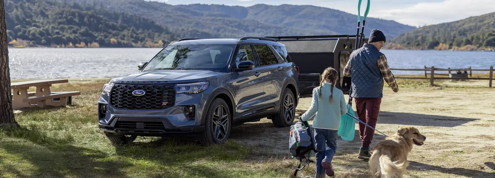 
A Ford Explorer is parked near a lake with a trailer attached to the back. A man, a girl, and a dog are walking away from the vehicle toward the right side of the frame. In the background are a lake and mountains.