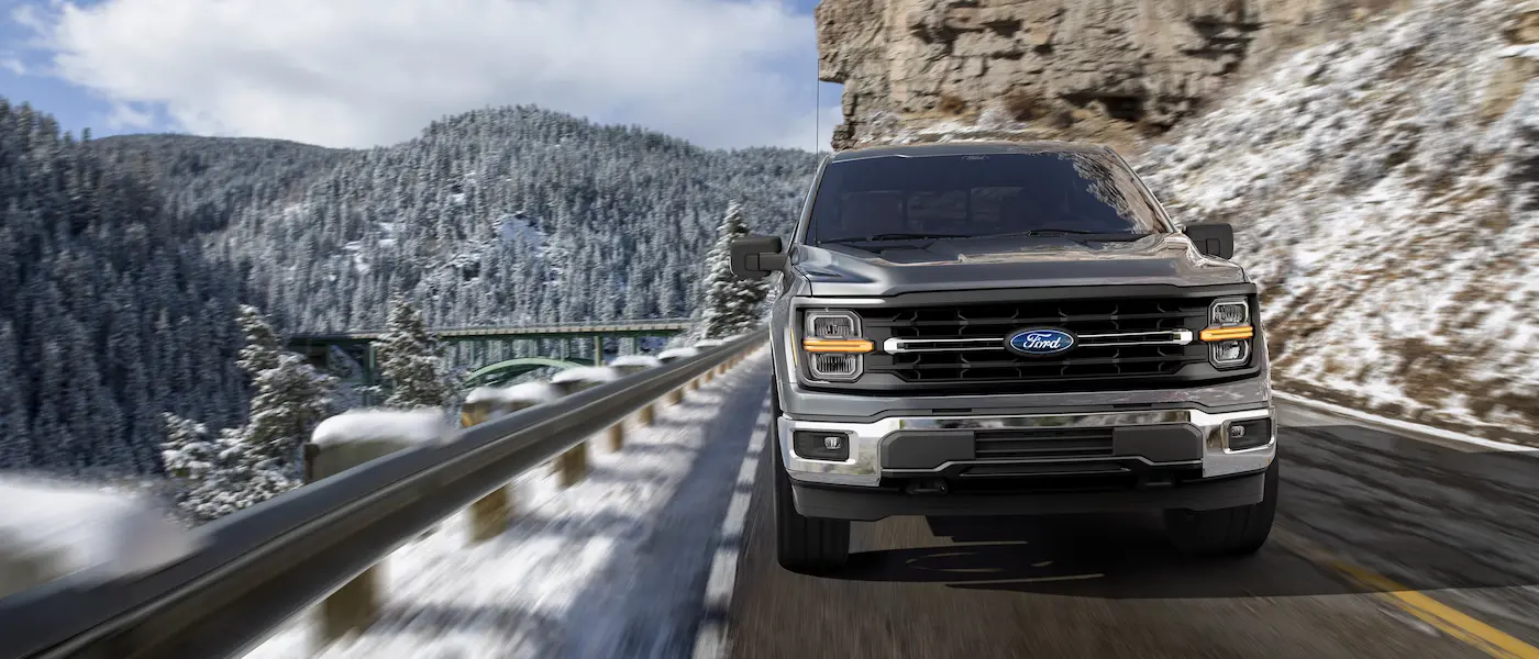 
A dark gray Ford F-150 pickup truck is driving on a snow-covered mountain road. The truck is centered and facing forward, with its headlights on. The background shows snow-covered pine trees, a bridge, and a rocky cliff face.