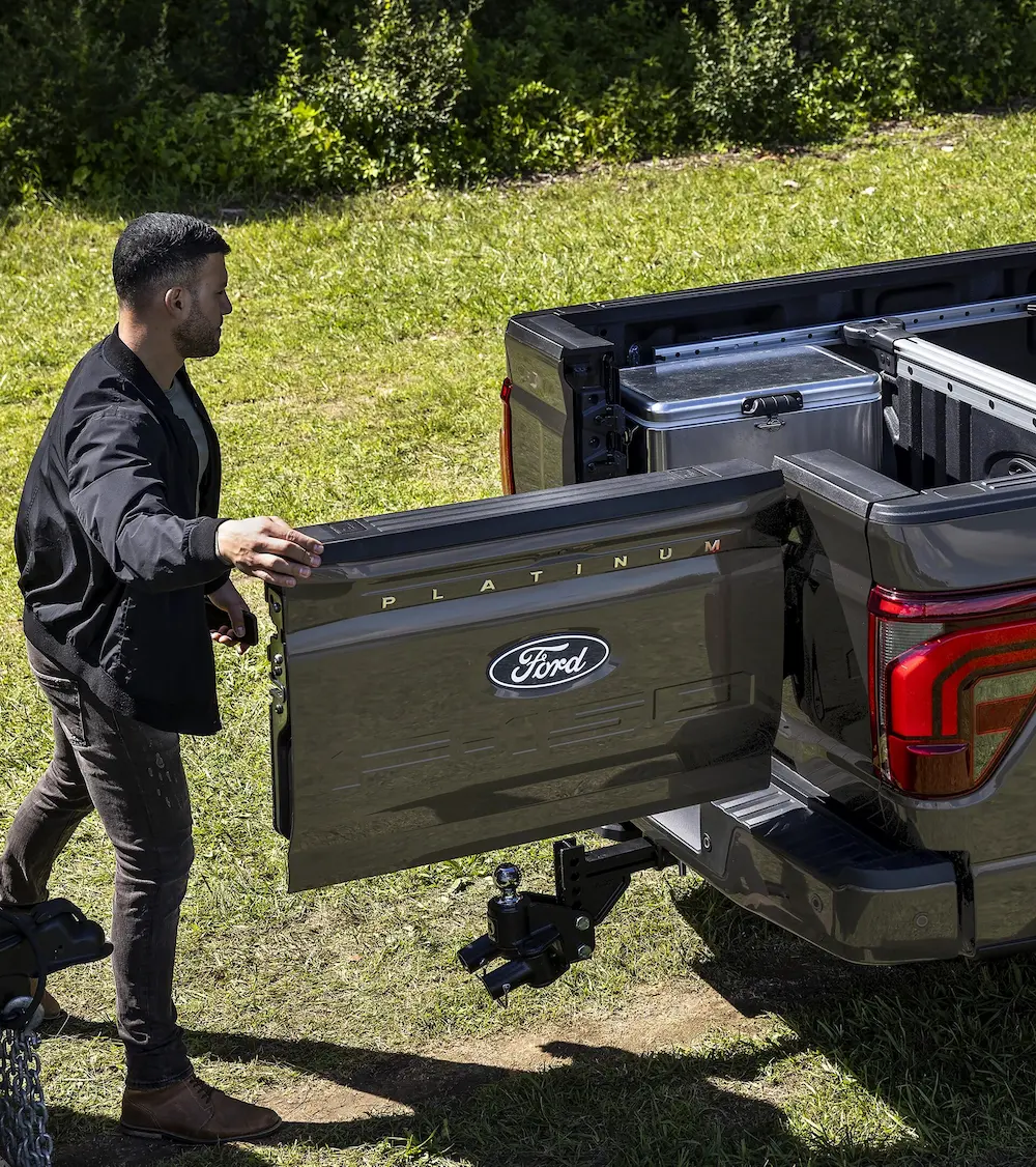 A man is standing on the left side of a dark gray Ford F-150 Platinum pickup truck, opening the multi-function tailgate. The truck bed contains a large metal toolbox. The background is a grassy field.