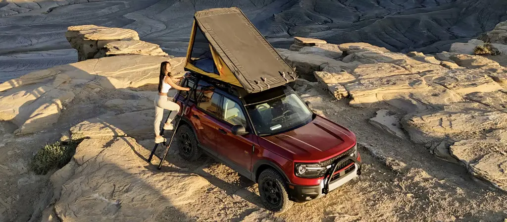 A woman climbs a ladder to a rooftop tent on a red Ford Bronco Sport