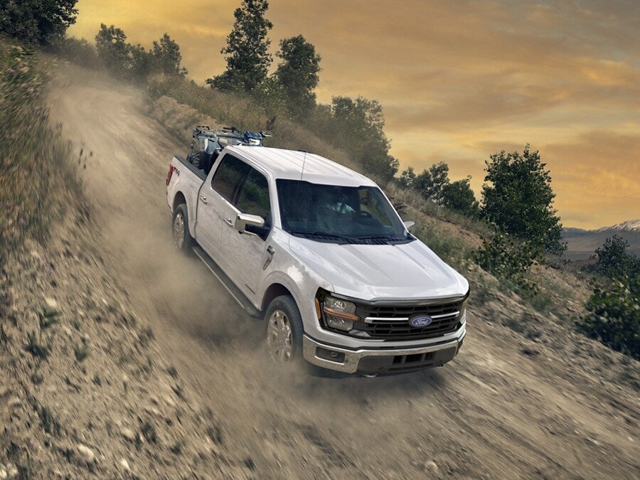 A white Ford F-150 pickup truck driving on a rough, dirt incline with a twilight sky and trees in the background.