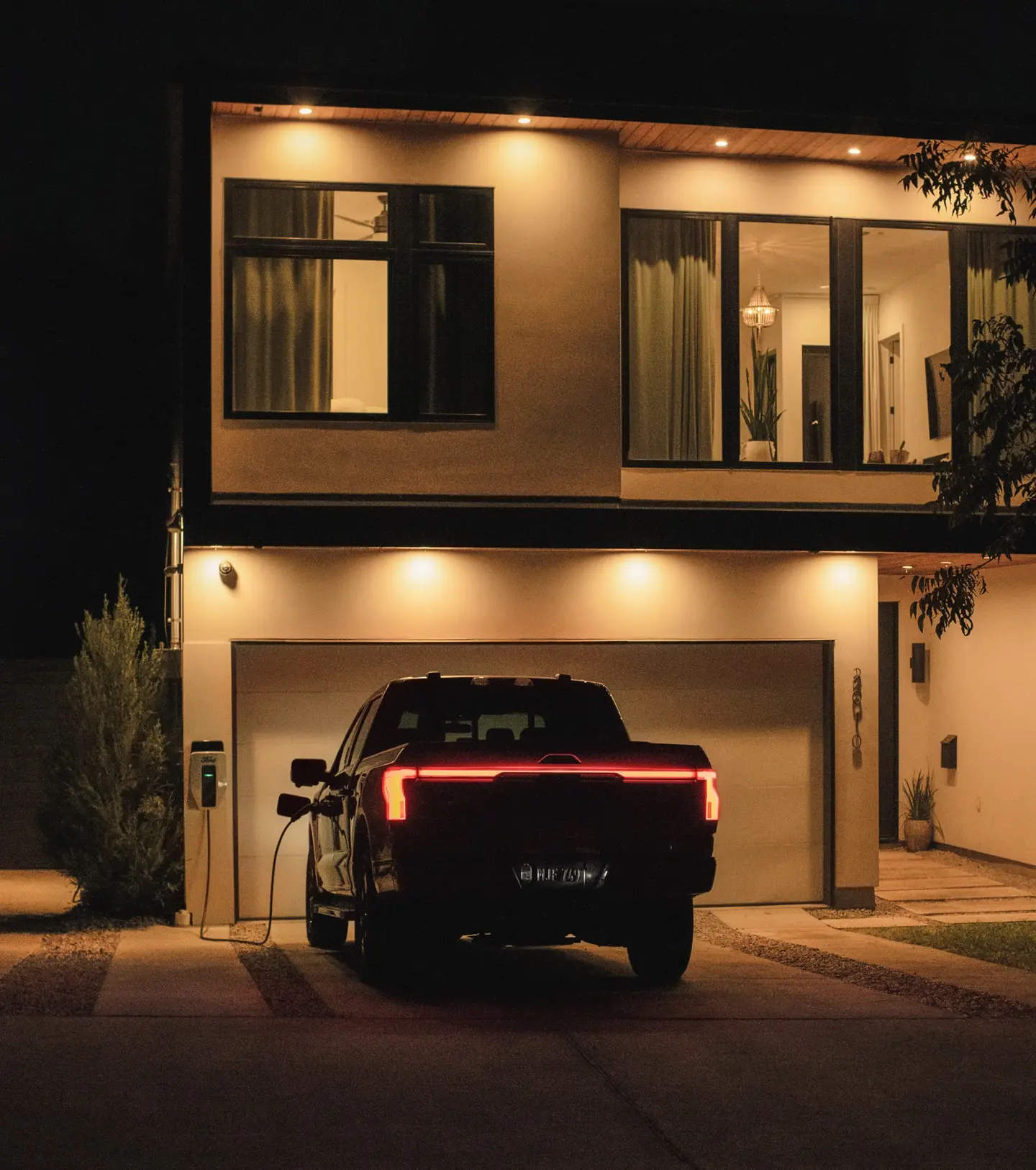 A Ford F-150 Lightning pickup truck is parked in a driveway at night, plugged into a charging station. 