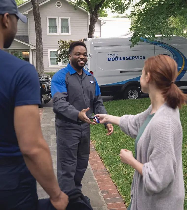 A woman is handing a key fob to a smiling Ford service technician. A Ford Mobile Service van is parked behind them.