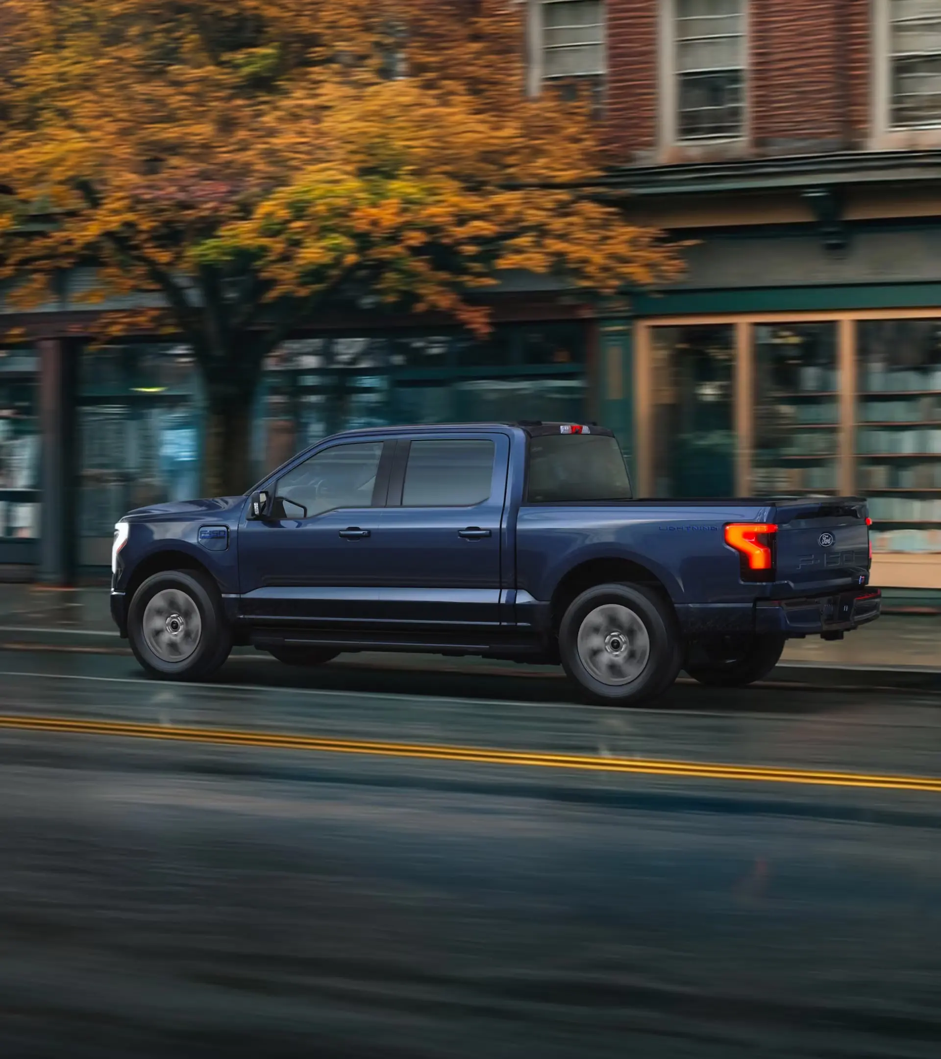 
A side view of a blue Ford F-150 Lightning electric pickup truck driving down a city street, with blurred buildings and trees in the background.