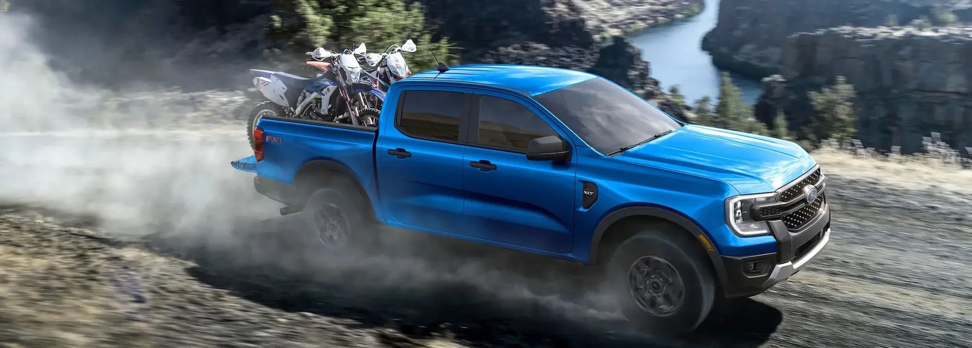 A blue Ford Ranger Raptor pickup truck, carrying two dirt bikes, is speeding down a dirt road.